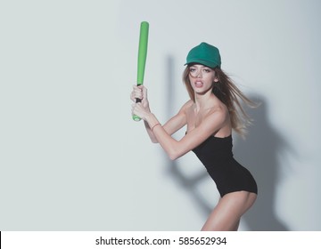 young pretty woman or cute sexy girl with beautiful long hair and adorable face in fashionable black bodysuit and cap holds green baseball bat or racket on grey background, copy space