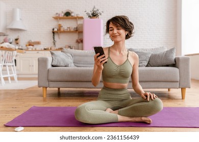 young pretty woman doing yoga at home wearing stylish sport outfit watching exercises on phone, meditation on mat, listening to music in earpods - Powered by Shutterstock