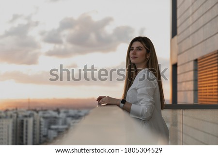 Young pretty woman dressed on white shirt looking at city from balcony, enjoying the sunset at terrace. Shot of beautiful young woman standing on the balcony and looking at the view. Relax, dreaming
