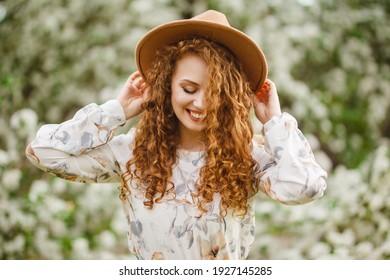 Young Pretty Woman Enjoys Standing Near Flowering Spring Tree. A Girl Wearing Beige Hat And White Dress Smiles Among Blooming Apple Trees. Spring Season Concept