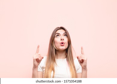 young pretty woman feeling awed and open mouthed pointing upwards with a shocked and surprised look against pink wall