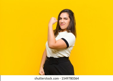 young pretty woman feeling happy, satisfied and powerful, flexing fit and muscular biceps, looking strong after the gym isolated against orange wall