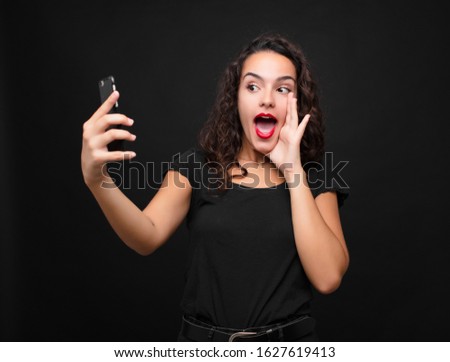 young pretty woman feeling happy, excited and positive, giving a big shout out with hands next to mouth, calling out holding a smartphone
