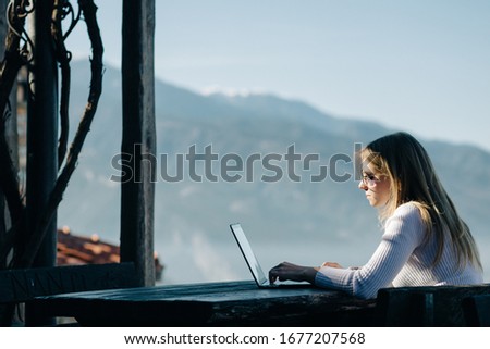 Young pretty woman freelancer works remotely at  laptop outside in front of spring Olympic mountains. Space for text