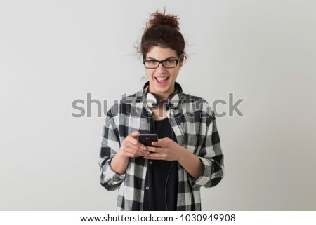 young pretty woman in glasses, using smartphone, emotional, laughing, positive, happy, headphones, isolated, checkered shirt, hipster style, student, typing message, looking in camera