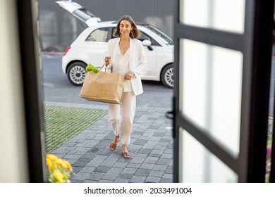 Young pretty woman going home with a shopping bag full of fresh groceries, coming home by a city car. View through the house door