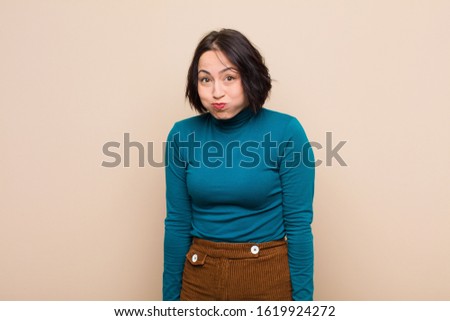 young pretty woman with a goofy, crazy, surprised expression, puffing cheeks, feeling stuffed, fat and full of food against beige wall