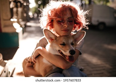 Young Pretty Woman In Grey Tee And Jeans Hugs Corgi. Charming Calm Girl In Sunglasses Plays With Dog Outside .