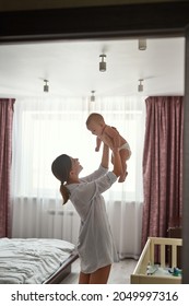 Young pretty woman holding up high cheerful little child on light window background, happy healthy family concept. Adorable mother and child in modern bedroom interior. Backlit photo.