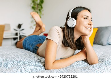 Young pretty woman listening music with wireless headphones while lying on bed. Millennial female relaxing at home enjoying online playlist. - Powered by Shutterstock