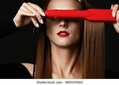 Young pretty woman with long silky straight hair in black clothes covering eyes with red hair straightener over dark background, close-up. Haircare, beauty, wellness, styling concept