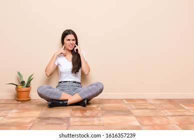 Young Pretty Woman Looking Angry, Stressed And Annoyed, Covering Both Ears To A Deafening Noise, Sound Or Loud Music Sitting On A Floor