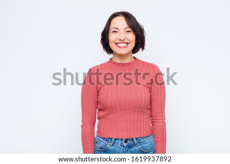 young pretty woman looking happy and goofy with a broad, fun, loony smile and eyes wide open against white wall
