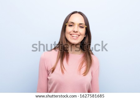 young pretty woman looking happy and goofy with a broad, fun, loony smile and eyes wide open against blue wall