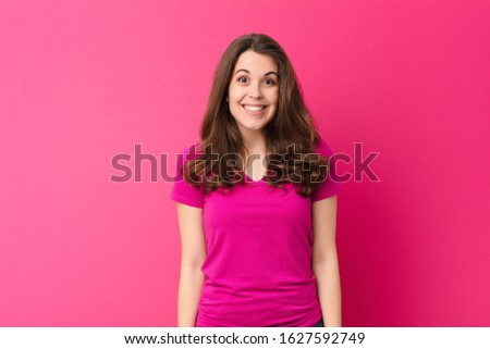 young pretty woman looking happy and goofy with a broad, fun, loony smile and eyes wide open against pink wall