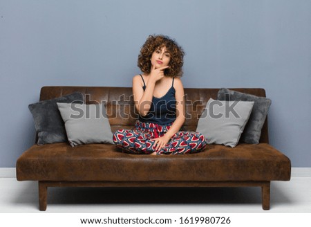 young pretty woman looking serious, thoughtful and distrustful, with one arm crossed and hand on chin, weighting options sitting on a sofa.