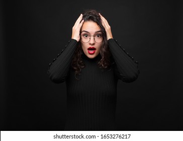young pretty woman looking unpleasantly shocked, scared or worried, mouth wide open and covering both ears with hands against black background.