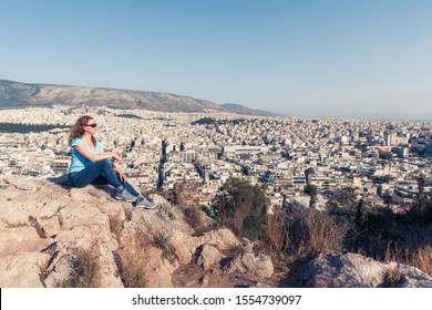 Young Pretty Woman Looks At Athens City, Greece. Beautiful Adult Girl Tourist Relaxes Overlooking Athens In Summer. Attractive Person Is On Background Of Urban Landscape. Travel And Vacation Concept.