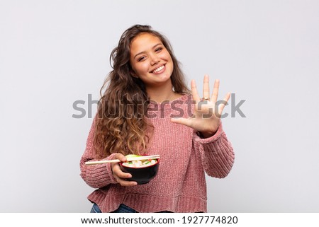 young pretty woman with noodles smiling and looking friendly, showing number five or fifth with hand forward, counting down