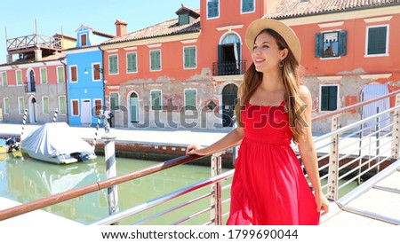 Young pretty woman in red dress walking on bridge in Burano village, Venice, Italy