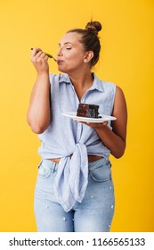 Young Pretty Woman In Shirt Happily Eating Chocolate Cake Over Yellow Background. Plus Size Model