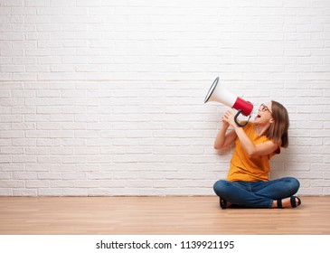 young pretty woman shouting on a megaphone sitting on wooden floor in front a brick wall