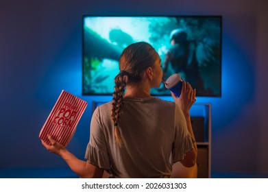 A young pretty woman sits in front of the TV with a pack of popcorn and a drink. Blue neon light. Color image. Close-up. Watching your favorite TV series, TV shows, weekend, relaxation. - Shutterstock ID 2026031303