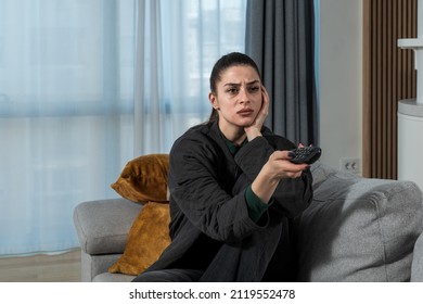 Young Pretty Woman Sitting At Home On The Lazy Weekend Watching Television Feeling Bored. Female In Cozy Apartment Enjoying Free Time While Her Boyfriend Or Husband Working.