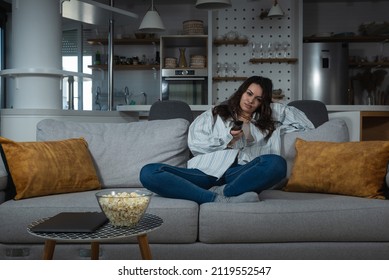 Young Pretty Woman Sitting At Home On The Lazy Weekend Watching Television Feeling Bored. Female In Cozy Apartment Enjoying Free Time While Her Boyfriend Or Husband Working.