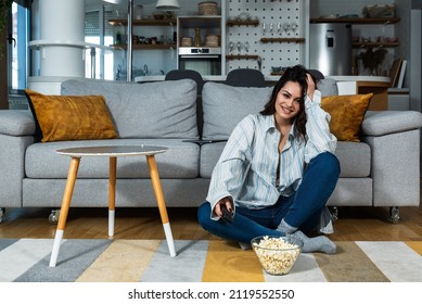 Young Pretty Woman Sitting At Home On The Lazy Weekend Watching Television Feeling Bored. Female In Cozy Apartment Enjoying Free Time While Her Boyfriend Or Husband Working.