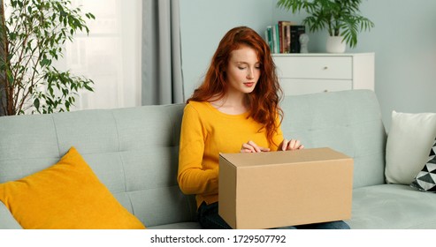 Young Pretty Woman Sitting On Couch In Living Room And Opening Carton Box. Cheerful Girl Getting Parcel At Home. Mail Delivery To House.