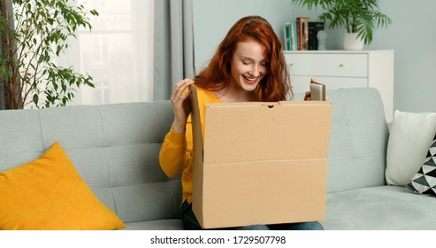 Young Pretty Woman Sitting On Couch In Living Room And Opening Carton Box. Cheerful Girl Getting Parcel At Home. Mail Delivery To House.