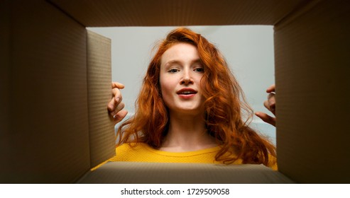 Young pretty woman sitting on couch in living room and opening carton box. Cheerful girl getting parcel at home. Mail delivery to house.
