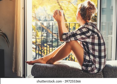 Young pretty woman  sitting at opened window drinking coffee and looking outside enjoys of rest
