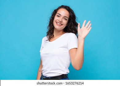 young pretty woman smiling happily and cheerfully, waving hand, welcoming and greeting you, or saying goodbye against blue background