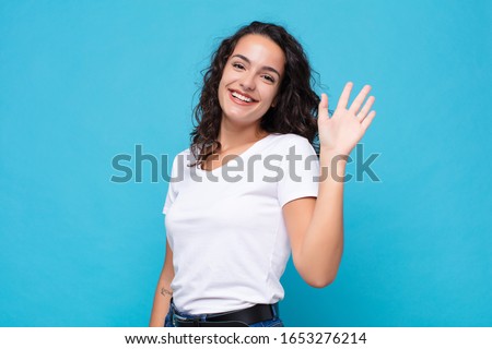 young pretty woman smiling happily and cheerfully, waving hand, welcoming and greeting you, or saying goodbye against blue background