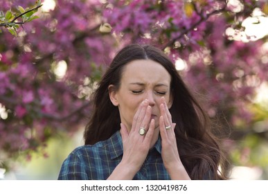 Young pretty woman sneezing in front of blooming tree. Spring allergy concept