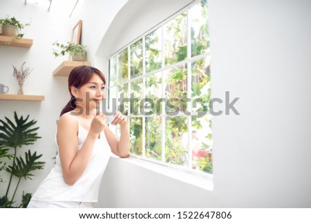 Young pretty woman standing behind window drinking coffee and looking outside enjoys of rest