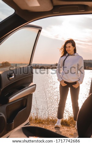 young pretty woman standing near car at river beach sunset on background