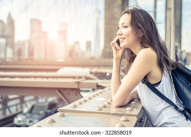 Young pretty woman talking at cellular phone on Brooklyn Bridge - Mixed race woman having a conversation at smartphone, city at sunset in the background - Powered by Shutterstock