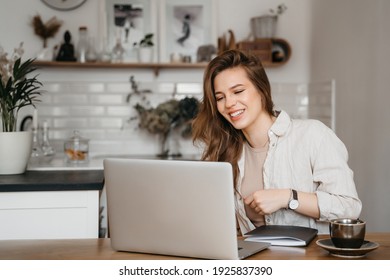 Young pretty woman talking on video call and waving hand while sitting at table inthe kitchen. High quality photo