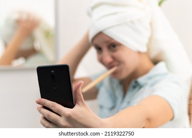 A young pretty woman with a towel on her head in a bright bathroom with a toothbrush and a smartphone in her hand brushes her teeth and communicates in social networks. Selective focus. Close-up - Powered by Shutterstock