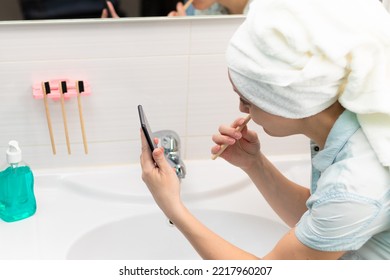 A young pretty woman with a towel on her head in a bright bathroom with a toothbrush and a smartphone in her hand brushes her teeth and communicates in social networks. Selective focus. Close-up - Powered by Shutterstock