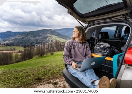 Young pretty woman traveler sitting in car trunk using laptop while traveling in mountains