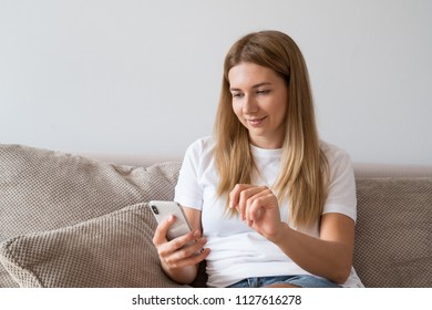 Young Pretty Woman Typing On Phone While Leaning On A Sofa. Girl Scrolling Smartphone.