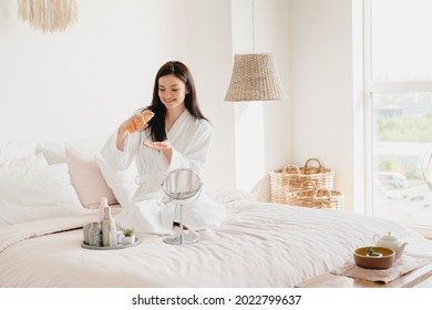 Young Pretty Woman Using Moisturizer Body Oil For Beauty Treatment, Skin Care. Girl In Spa Bathrobe Relaxing On Her Bed, Applying Cosmetics, Taking Care Of Her Hair And Nails