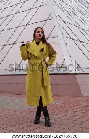 Young pretty woman walking on an urban street sidewalk along city building. She looks like an independent millennial female tourist wearing a colorful yellow coat traveling solo on foot.
