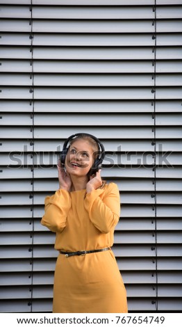 Young pretty woman wearing yellow dress standing against shutter and listening to music with headphones