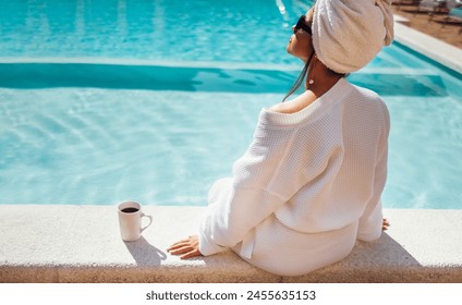 Young pretty woman in a white bathrobe by the swimming pool and holds a cup of coffee. Relaxed caucasian female in sunglasses and a towel on her head sits at the poolside. Vacation and spa concept. - Powered by Shutterstock