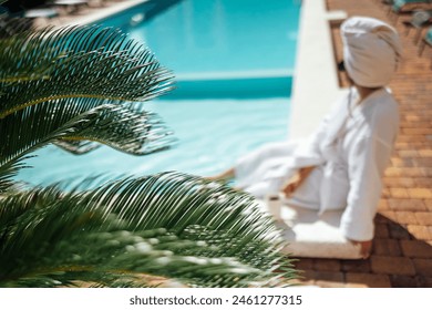 Young pretty woman in a white bathrobe by the swimming pool and holds a cup of coffee. Relaxed caucasian female in sunglasses and a towel on her head sits at the poolside. Vacation and spa concept. - Powered by Shutterstock